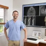 [LAST_NAME] standing and smiling in an exam room